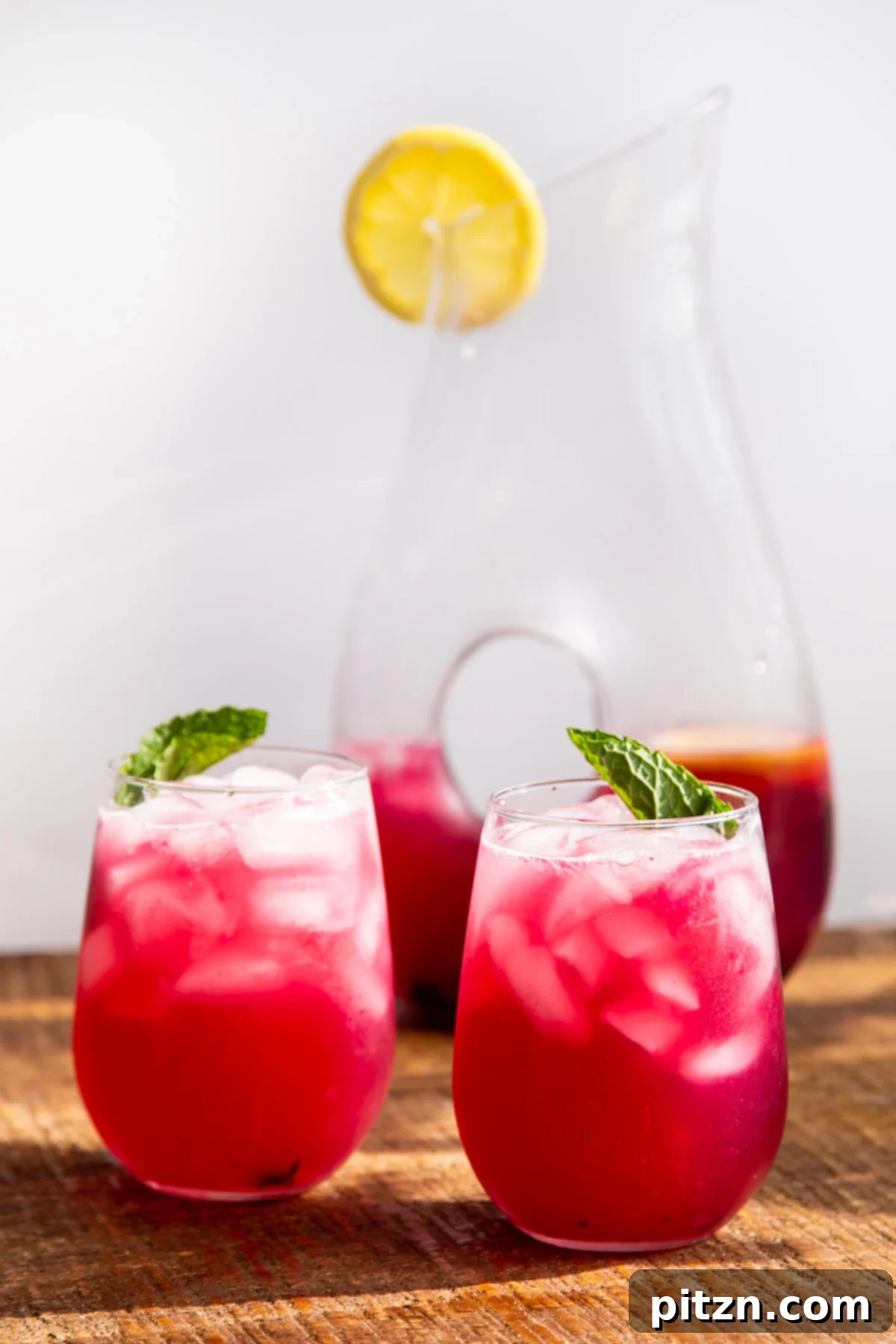 Two glasses of homemade fresh blueberry lemonade with a large jar of lemonade in the background, garnished with lemon slices and mint leaves.
