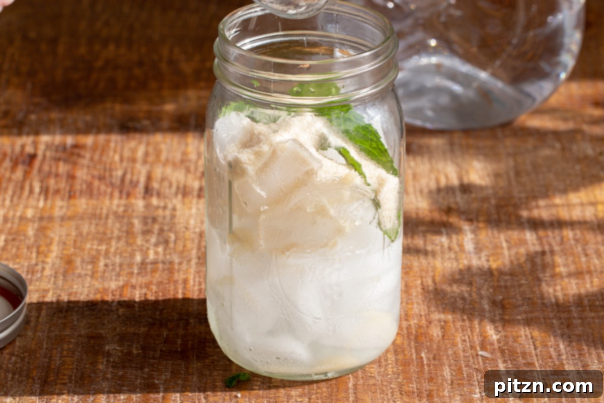 Ice cubes, lemon juice, and mint leaves are already placed in a mason jar, awaiting the muddled blueberry mixture.