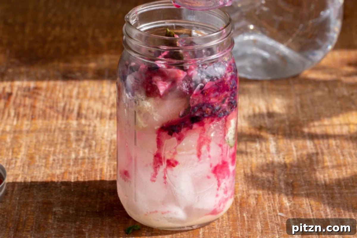 Muddled blueberries are being added to a mason jar already containing ice cubes and lemon juice, starting the lemonade assembly.
