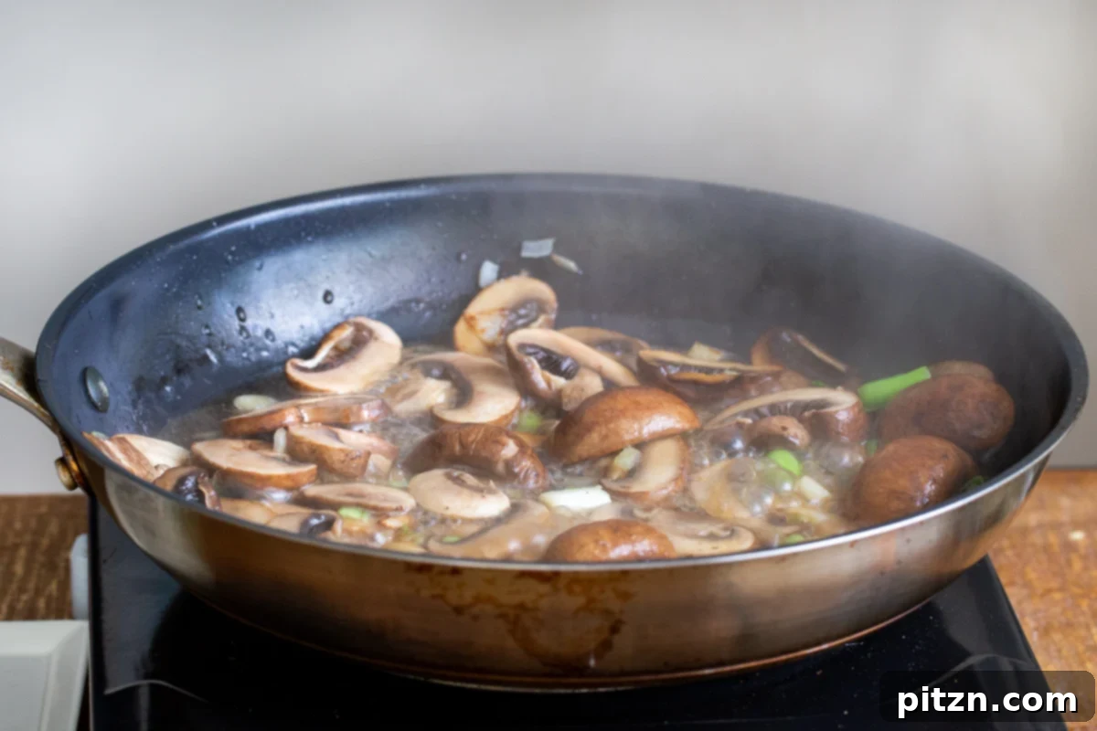 Sautéed mushrooms simmering in a black pan with broth, absorbing flavors and softening.