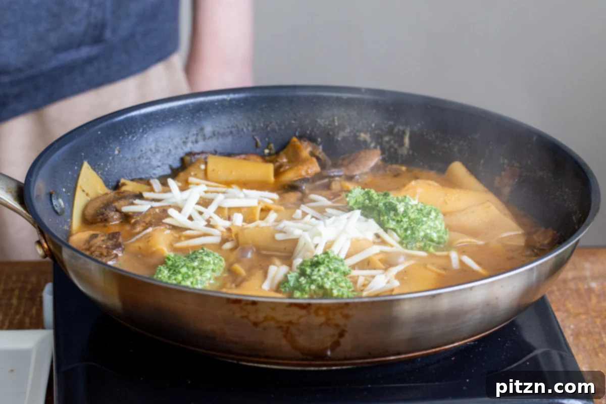 Lasagna pasta simmering gently in a skillet, adorned with dollops of vibrant pesto and melting vegan cheese, creating a rich texture.