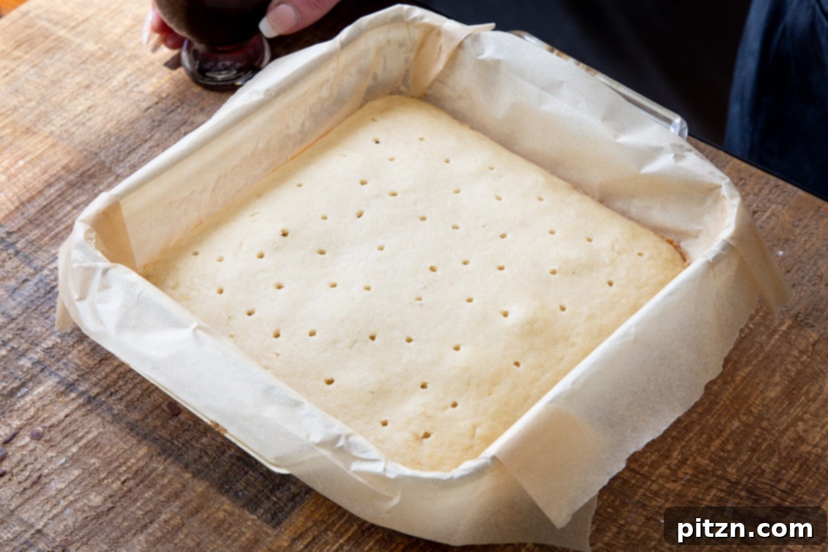 Plant-Based Tiramisu Tray Bake 13 A freshly baked vegan sponge cake in a square pan being systematically poked with holes using a fork, preparing it for coffee syrup.