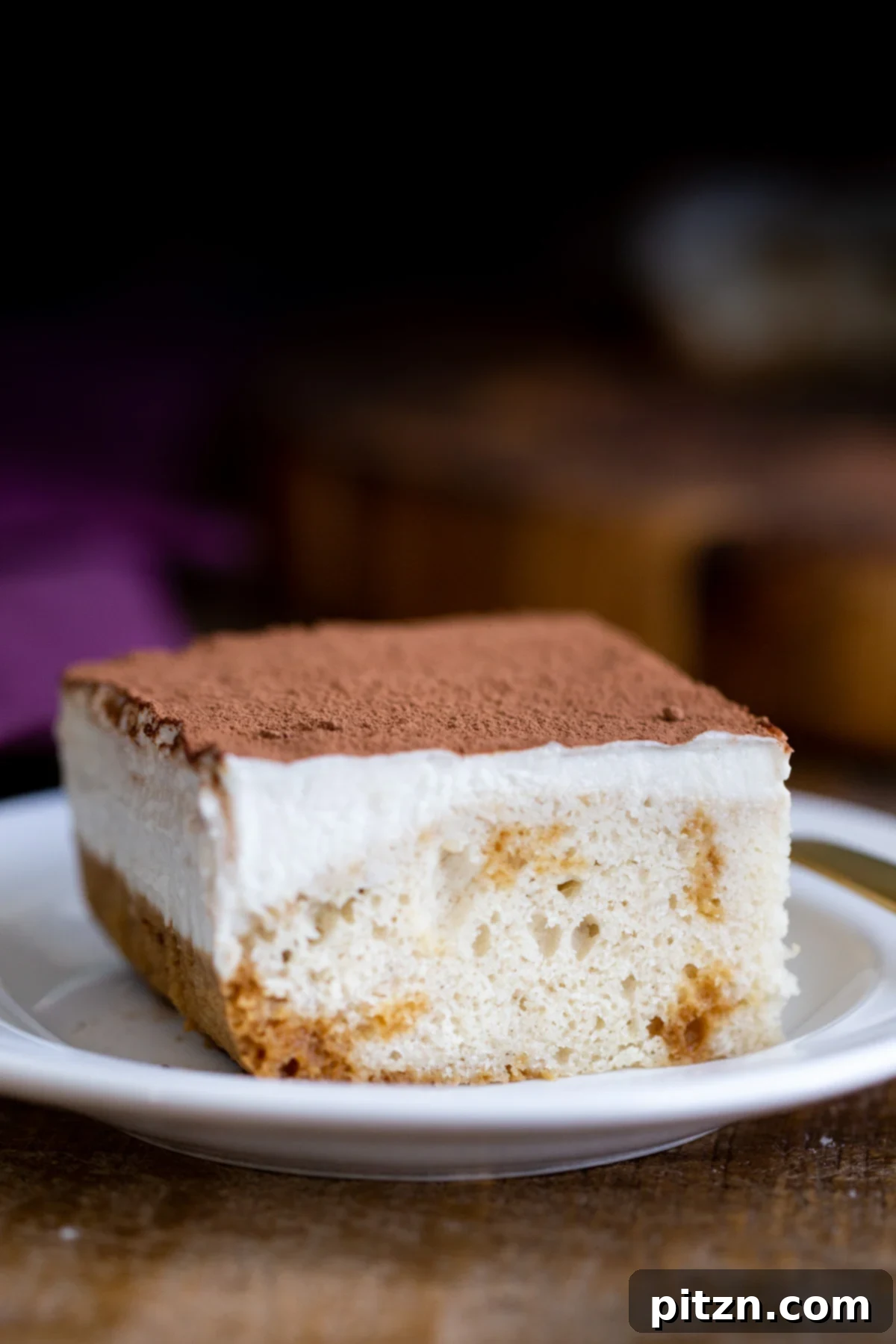 Plant-Based Tiramisu Tray Bake 5 A square piece of vegan tiramisu sheet cake, delicately dusted with cocoa powder, poised on a small white plate, highlighting its elegant layers.