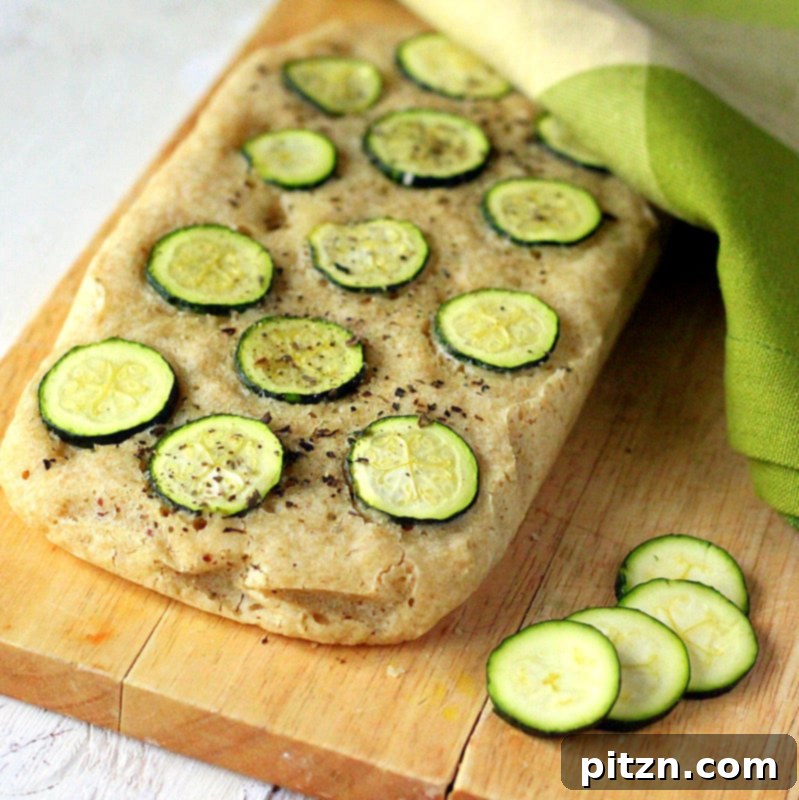 Naturally Gum-Free Vegan Gluten-Free Zucchini Basil Focaccia 2 A golden-brown Gluten-free Zucchini Basil Focaccia, topped with green zucchini slices and fresh basil leaves, resting on a rustic wooden board. The bread has a beautiful texture and a scattering of herbs.