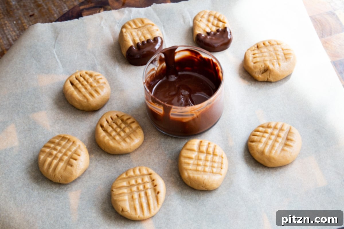 A sheet of parchment paper laid out with shaped no-bake peanut butter cookies, positioned beside a small bowl of glistening melted chocolate for dipping.