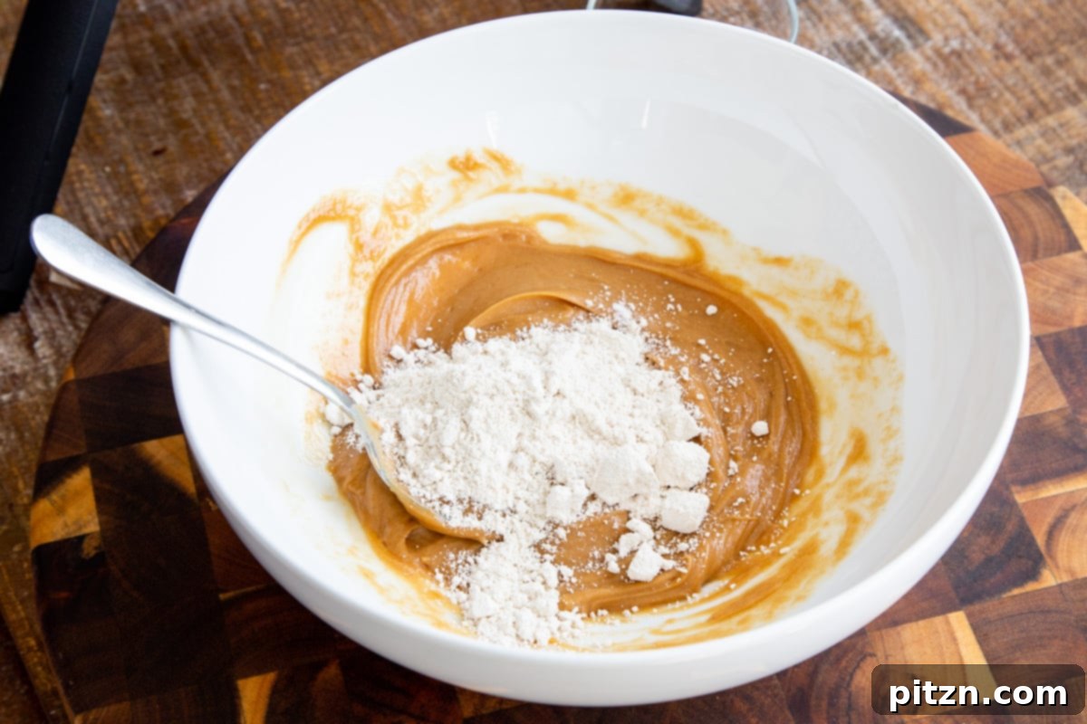 Gluten-free flour being carefully stirred into the smooth peanut butter mixture in a white bowl, creating the cookie dough.