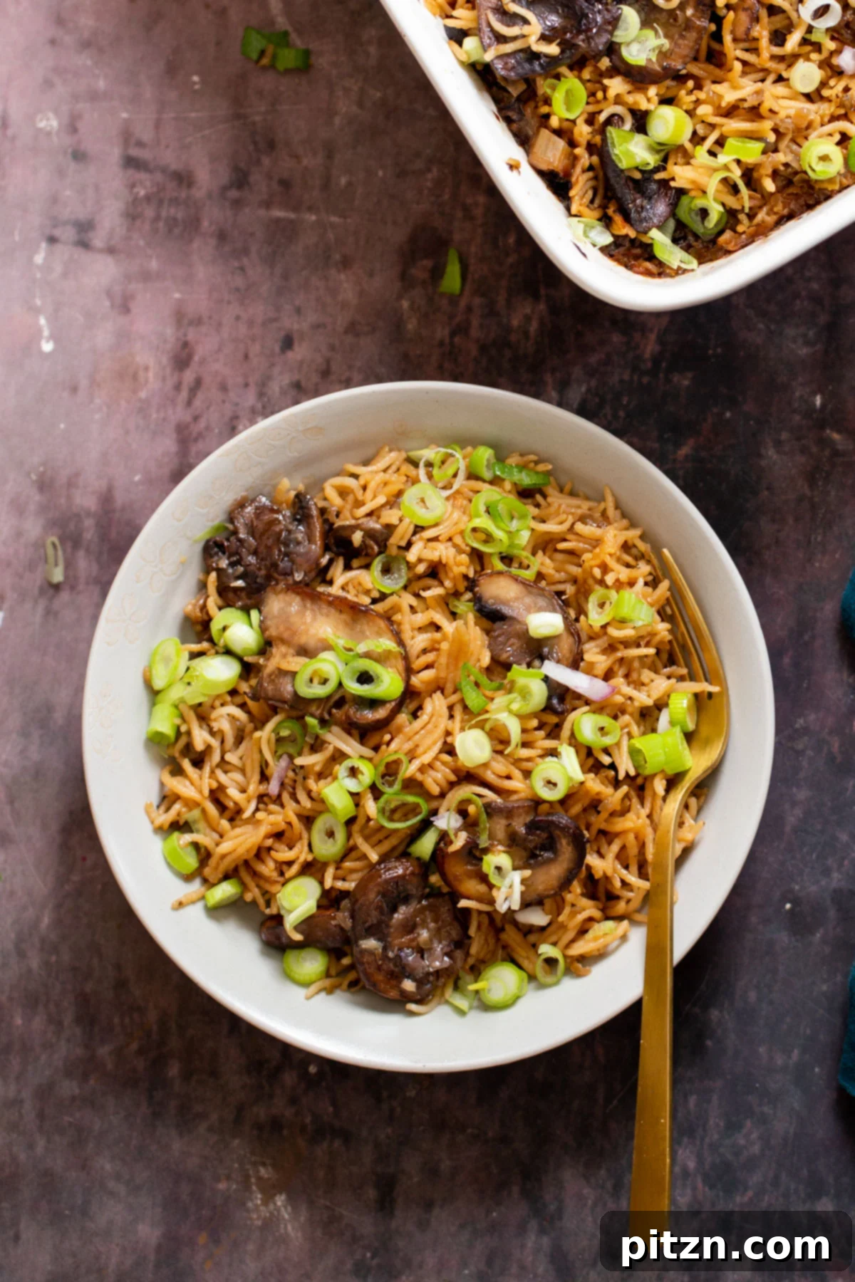 A golden fork rests on the side of a plate and a casserole dish, both showcasing the fluffy baked mushroom rice.