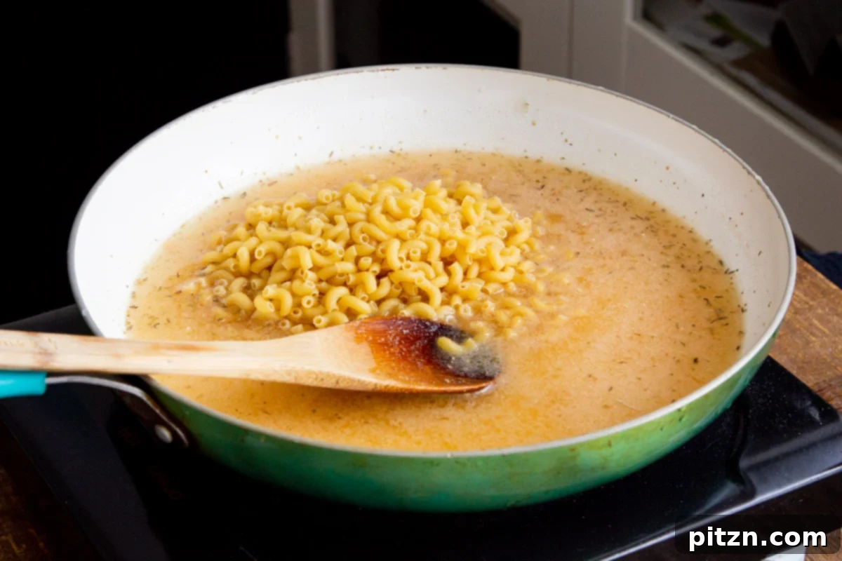 Fiery Vegan Skillet Mac and Cheese 9 Dried elbow pasta being carefully added to a skillet filled with aromatic, seasoned broth on the stovetop.