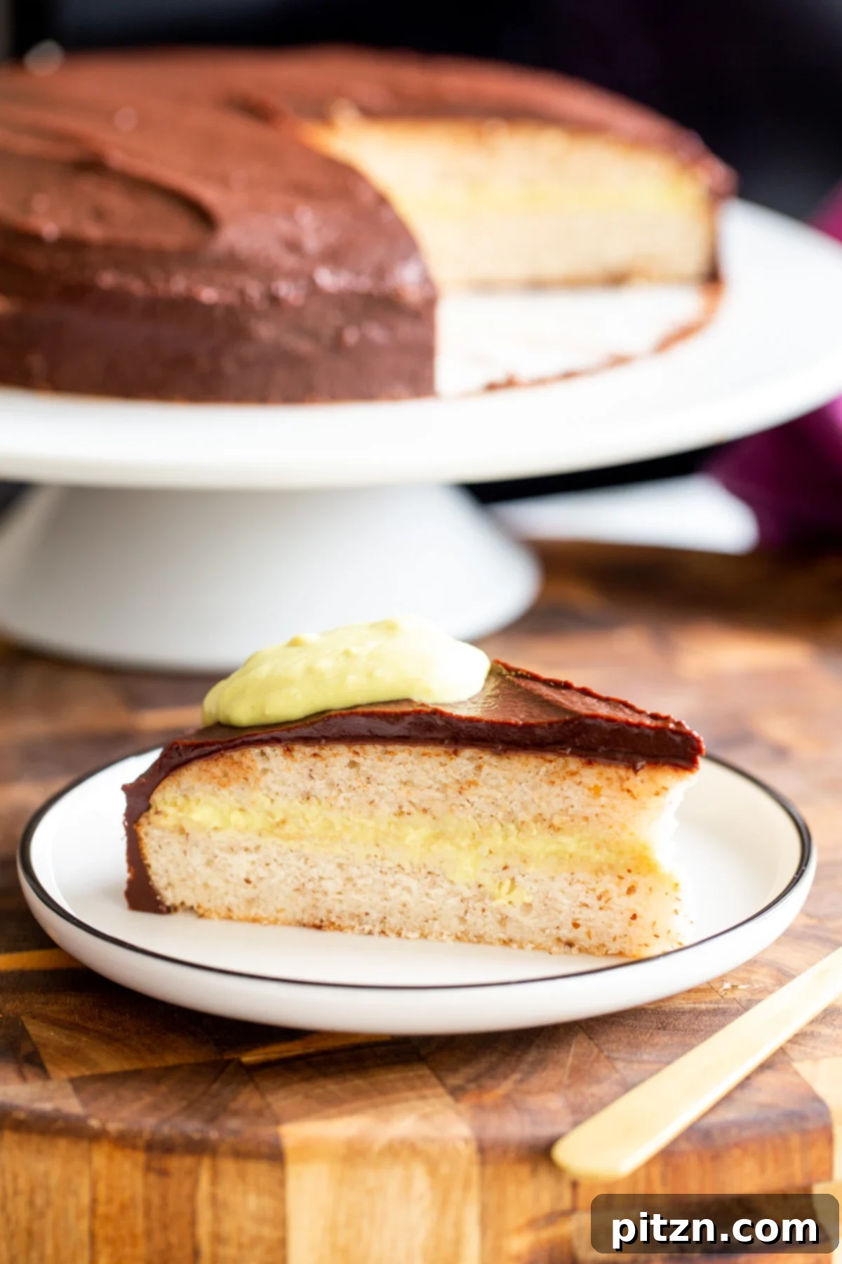 Plant-Based Boston Dream Cake 2 A perfectly sliced piece of Vegan Boston Cream Pie Cake revealing its layers, with the full cake visible in the softly blurred background.