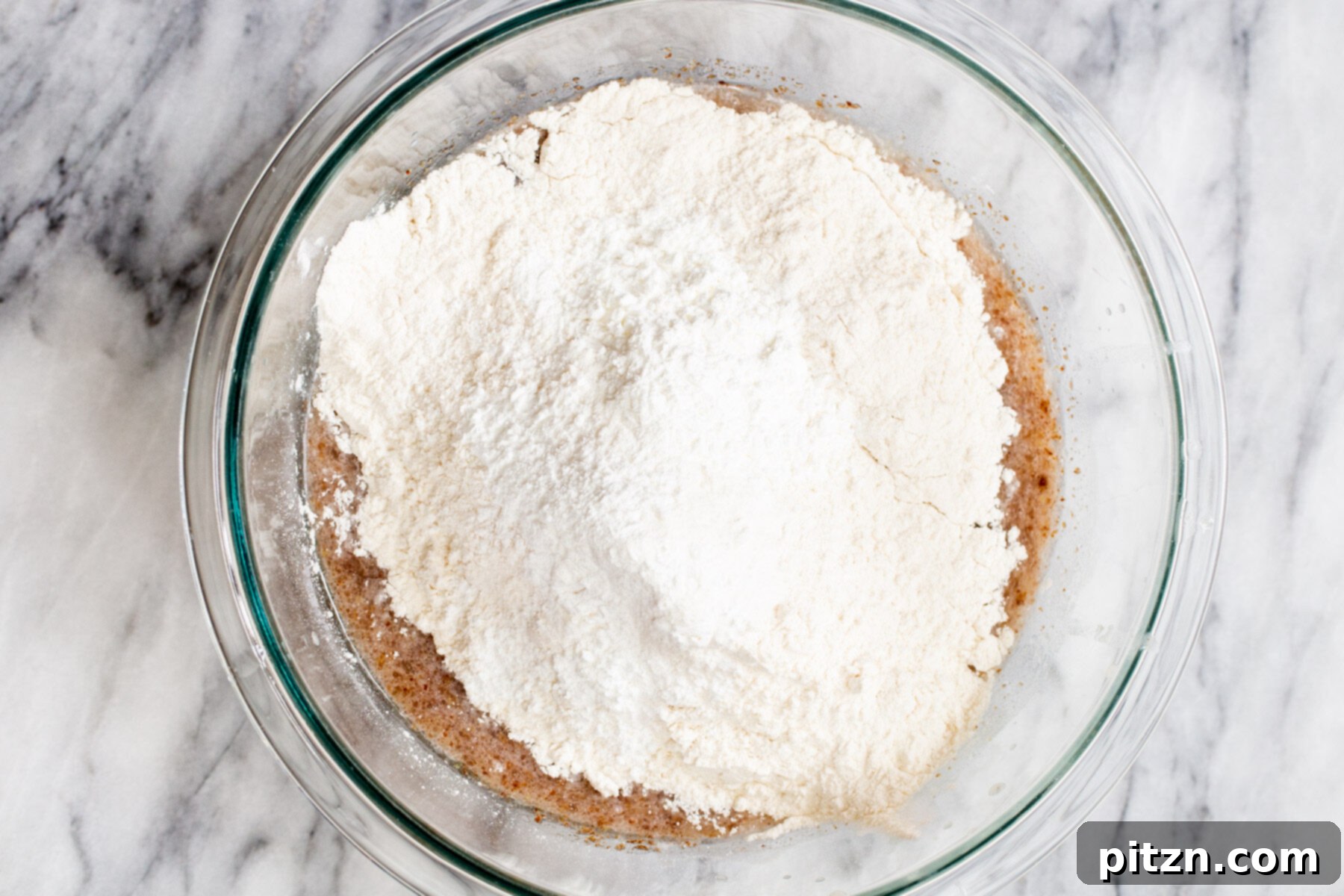 Plant-Based Boston Dream Cake 10 Dry ingredients like flour, flax meal, cornstarch, baking powder, and salt layered on top of the wet ingredients in a glass bowl for the vegan cake batter.