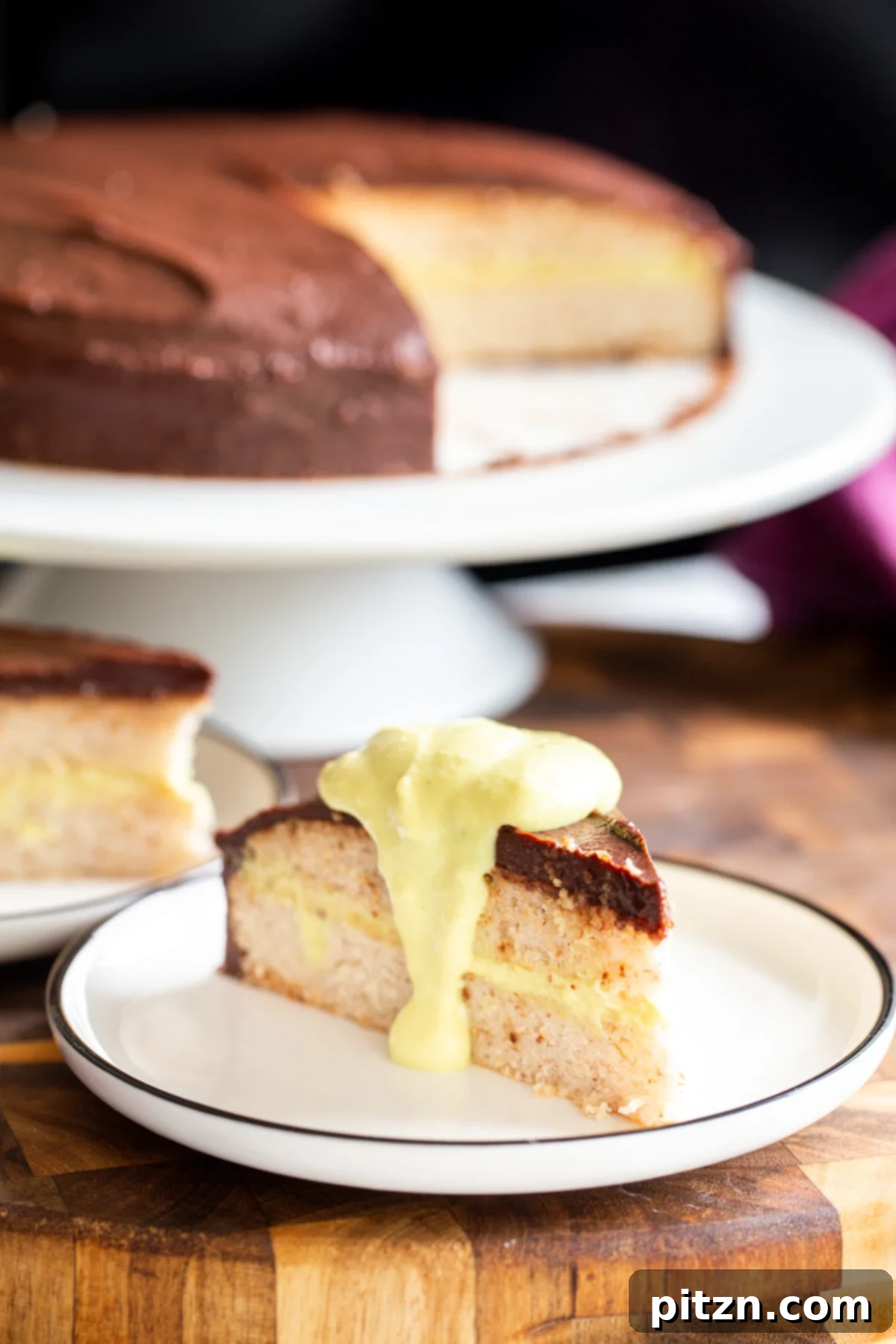 Plant-Based Boston Dream Cake 4 A generous slice of vegan Boston Cream Pie Cake on a plate, with the remaining cake proudly displayed on a cake stand in the background, ready for more servings.
