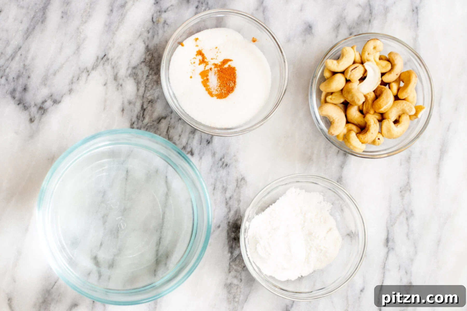 Plant-Based Boston Dream Cake 6 Ingredients for making vegan vanilla custard, including raw cashews, water, vanilla extract, almond extract, turmeric, sugar, and thickeners, arranged on a marble countertop.