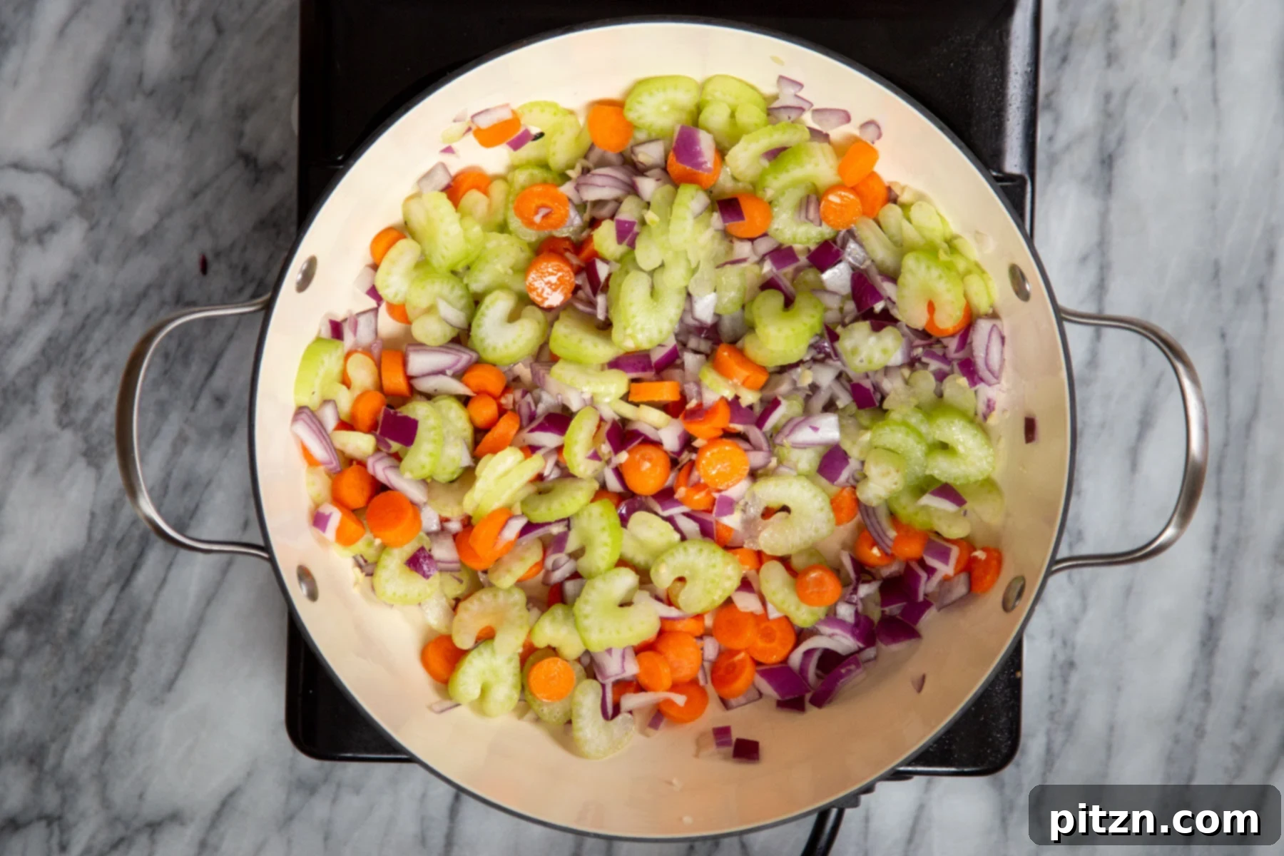 Carrots, onions, and celery glistening in a sauté pan, having softened and developed rich colors.