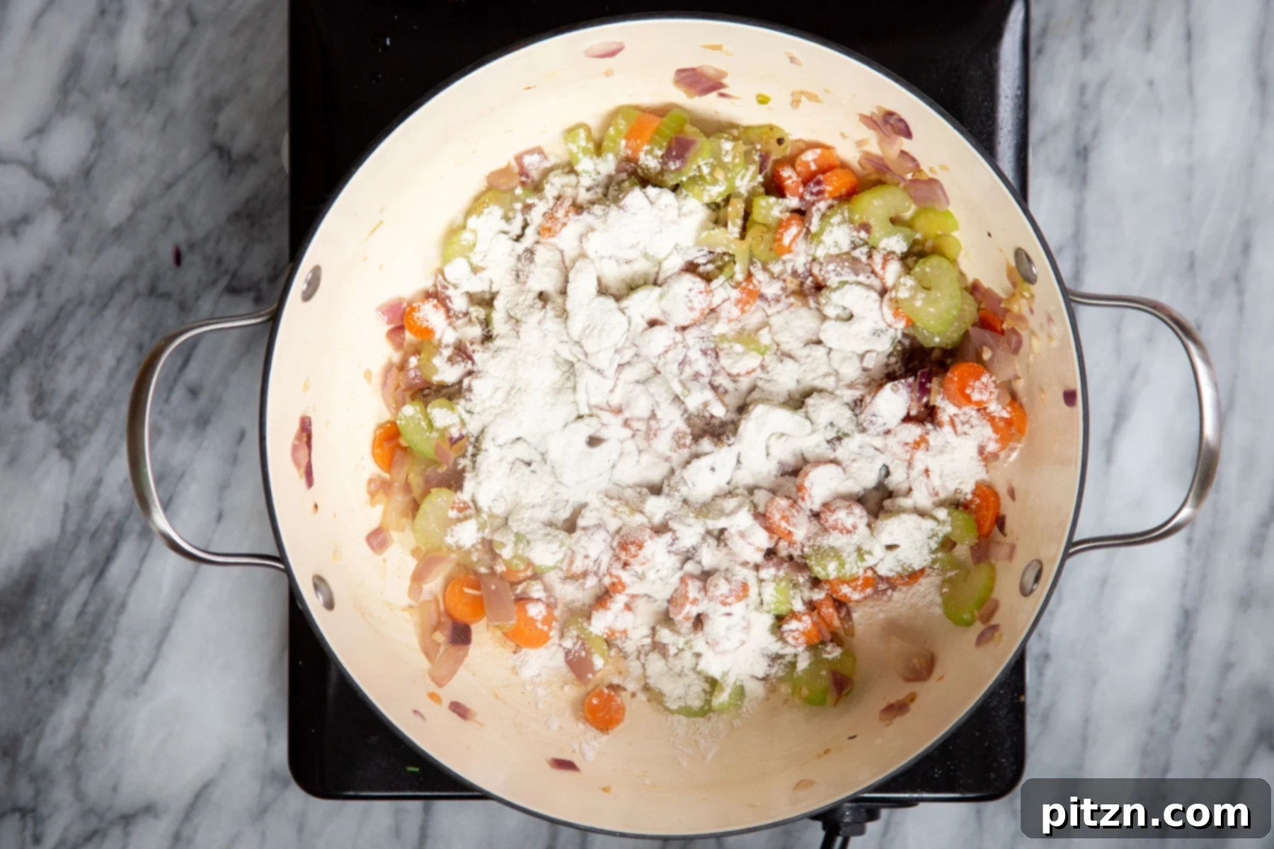Mixed vegetables and flour being stirred in a saucepan on a cooking hob, preparing for the liquid additions.