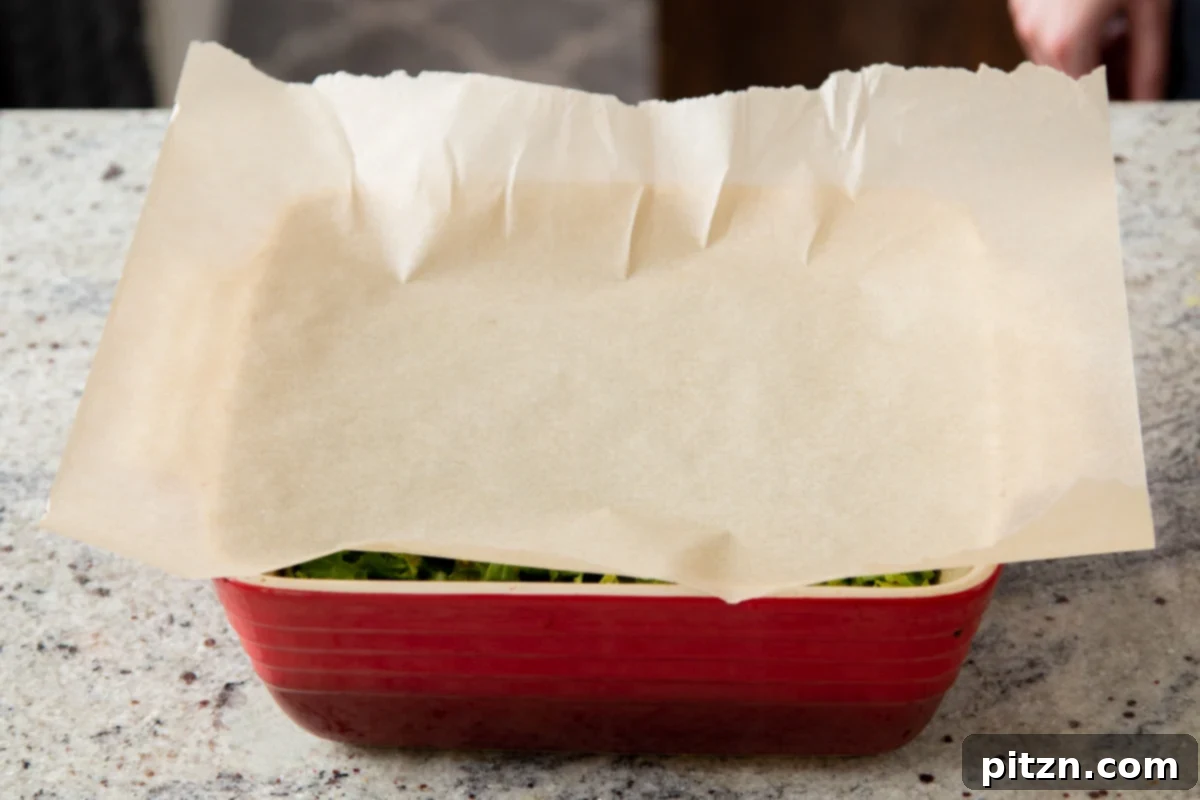A view of mustard greens simmering in a red casserole dish, covered with parchment paper for baking.