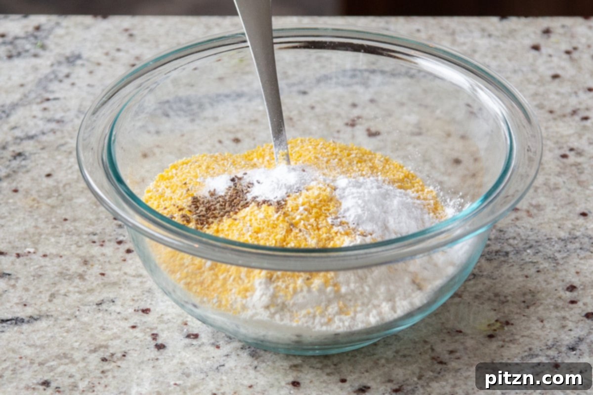 Dry ingredients for cornbread topping, including cornmeal, flour, and various spices, mixed in a clear glass bowl.