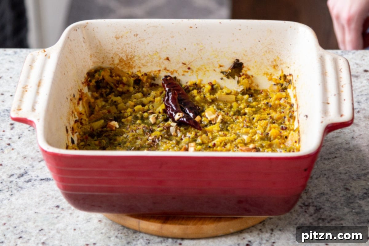 Tempered spices, including chili and garlic, being gently mixed into the cooked mustard greens in a red casserole dish.