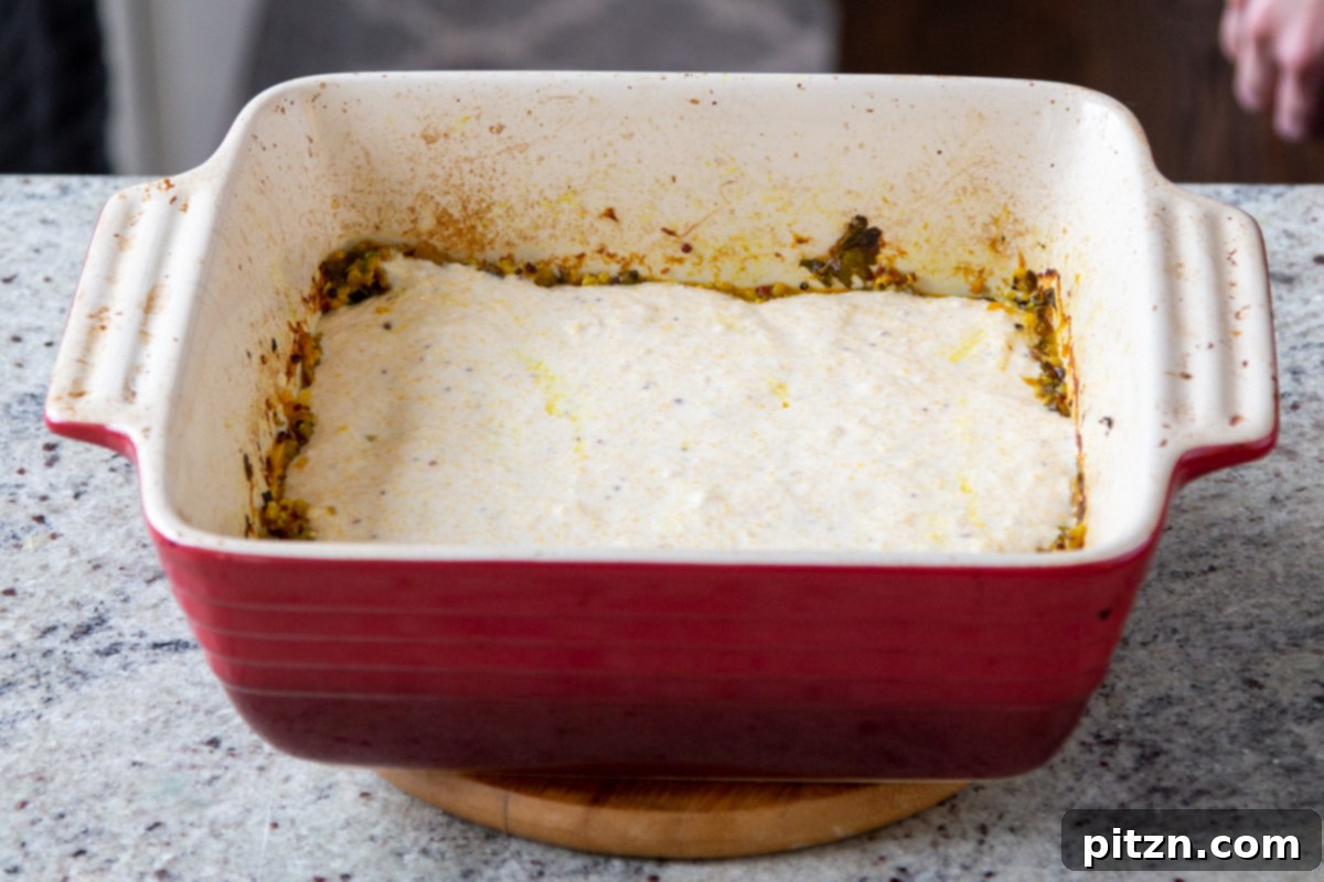 The cornbread topping batter being spread evenly over the spiced saag greens in a red casserole dish.