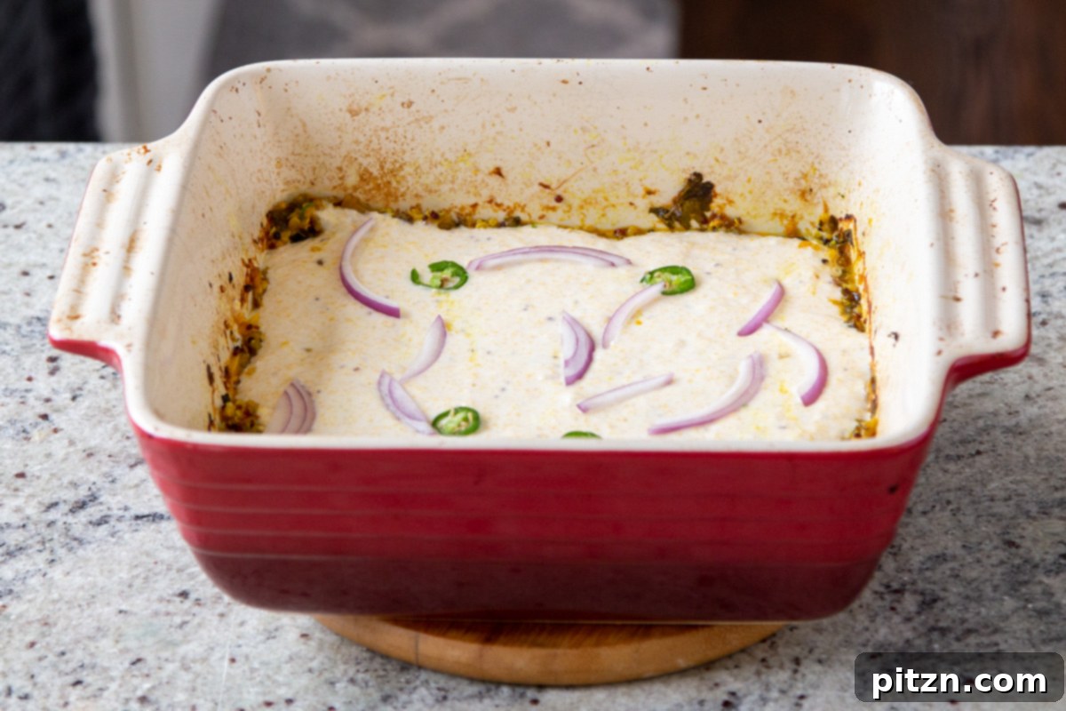 Saag casserole topped with a golden cornbread layer, adorned with sliced onions and chilies before the final bake.