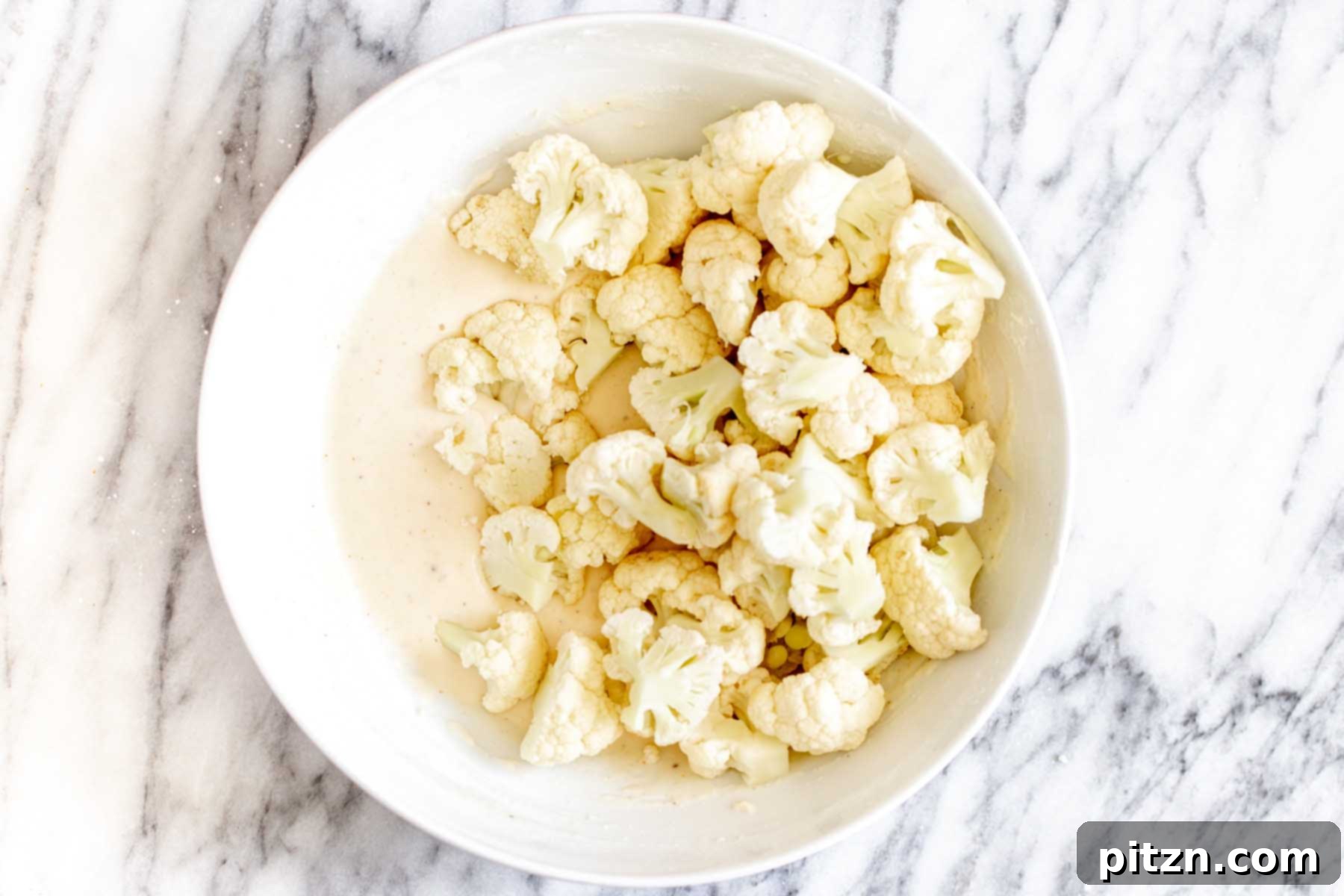 Cauliflower florets placed into a bowl of batter, ready to be tossed and coated for baking.