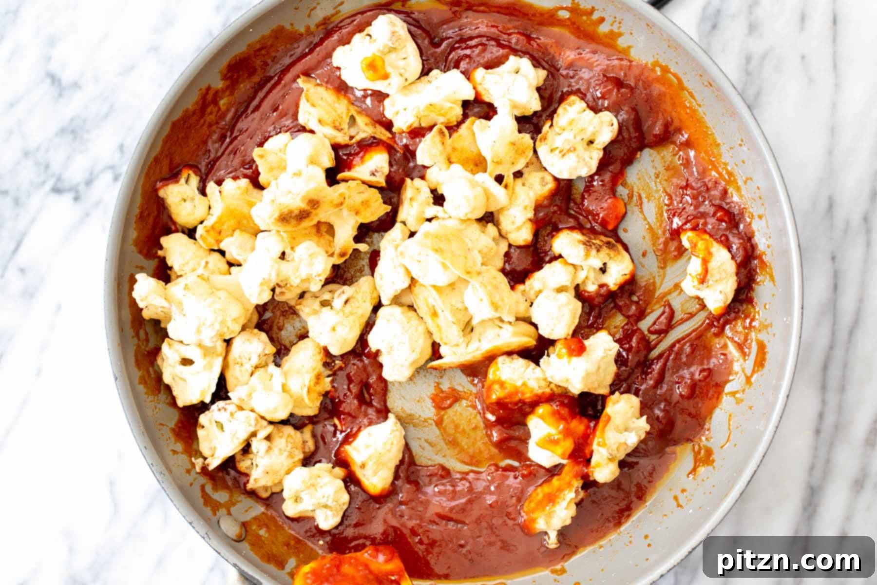 Baked cauliflower florets being added to a skillet containing the cooked gochujang sauce, ready for tossing.
