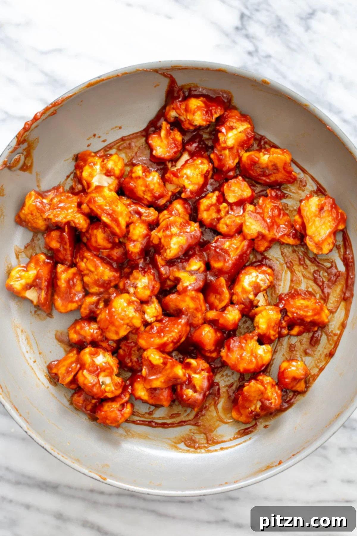 Freshly baked cauliflower florets being tossed in a glistening, thick gochujang sauce in a large skillet, ensuring every piece is thoroughly coated.
