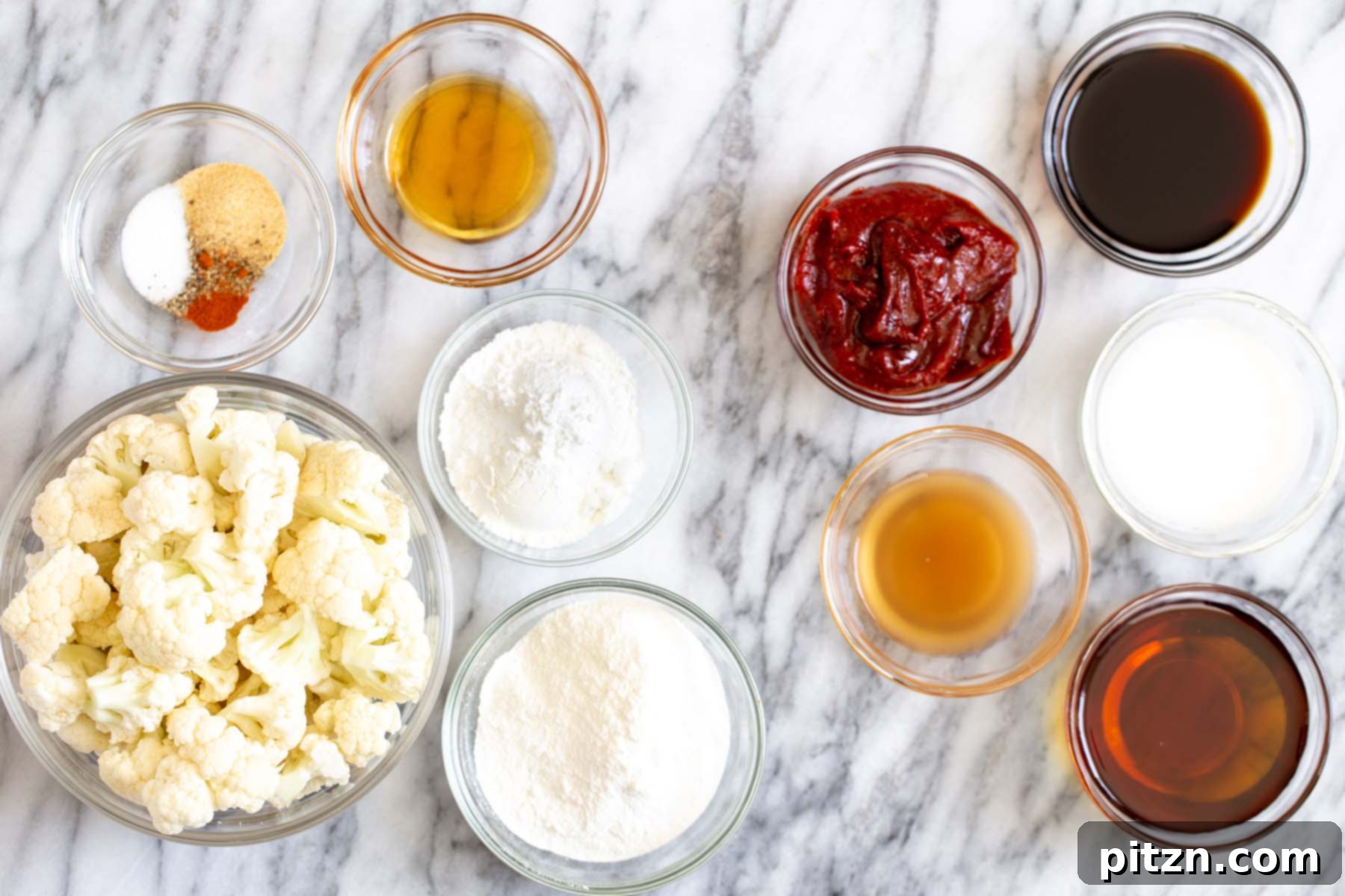 Various gochujang cauliflower ingredients, including flours, spices, oils, and the gochujang paste, arranged in small bowls on a elegant marble table.
