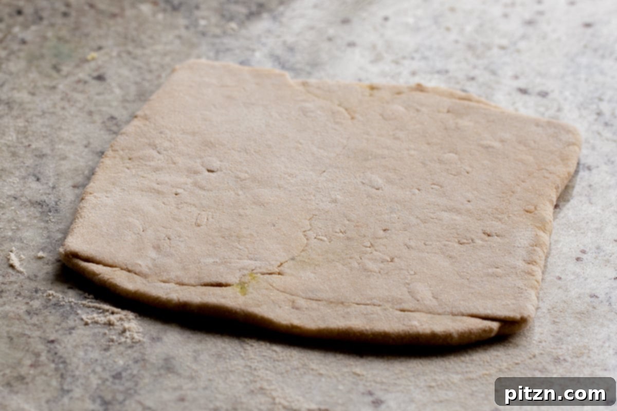 A stuffed aloo paratha, perfectly rolled into a square shape, positioned on a floured surface and ready for cooking.