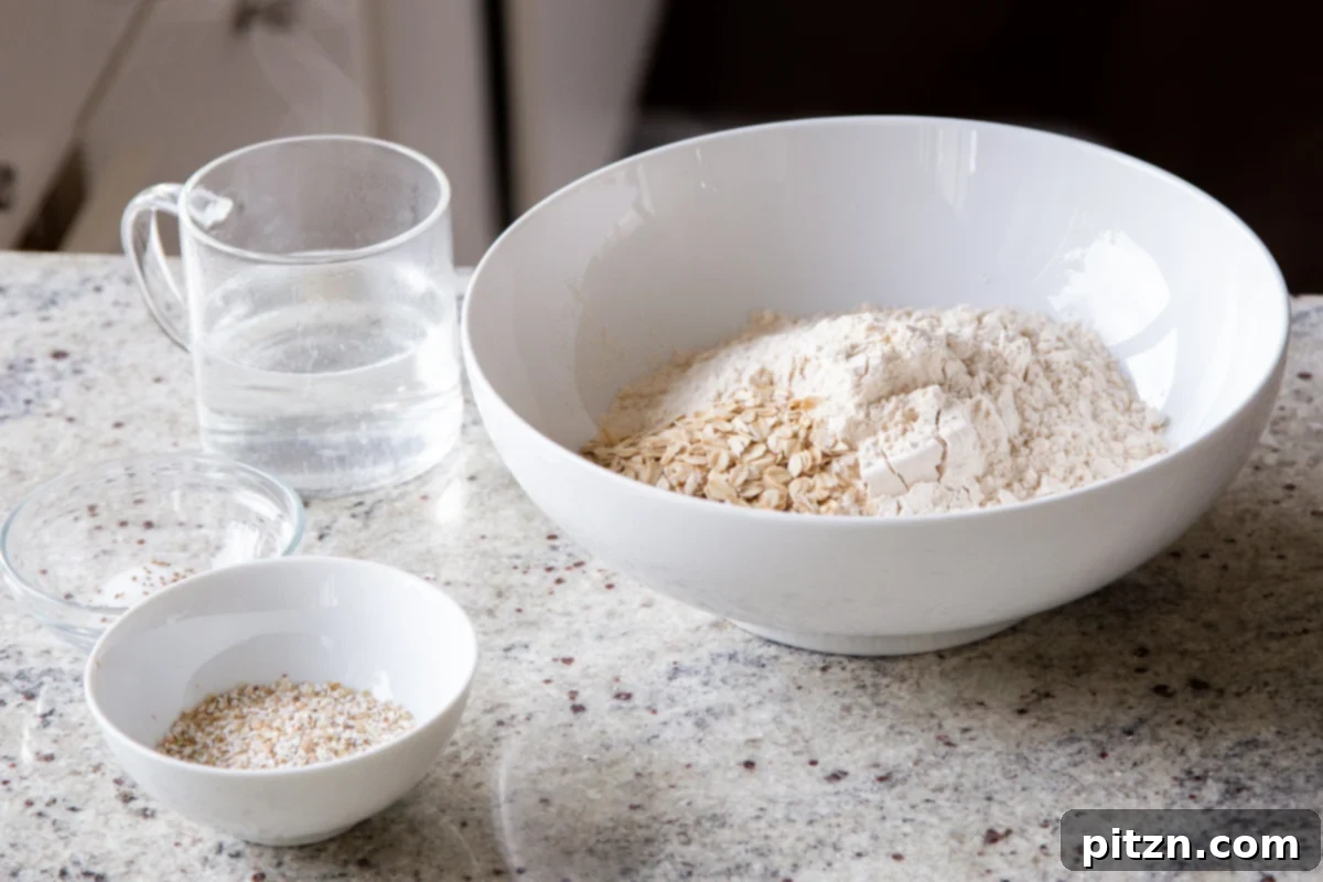 A white mixing bowl contains a blend of chapati flour and whole grains, with a separate small bowl of buckwheat groats and a cup of warm water nearby on a clean countertop, ready for dough preparation.