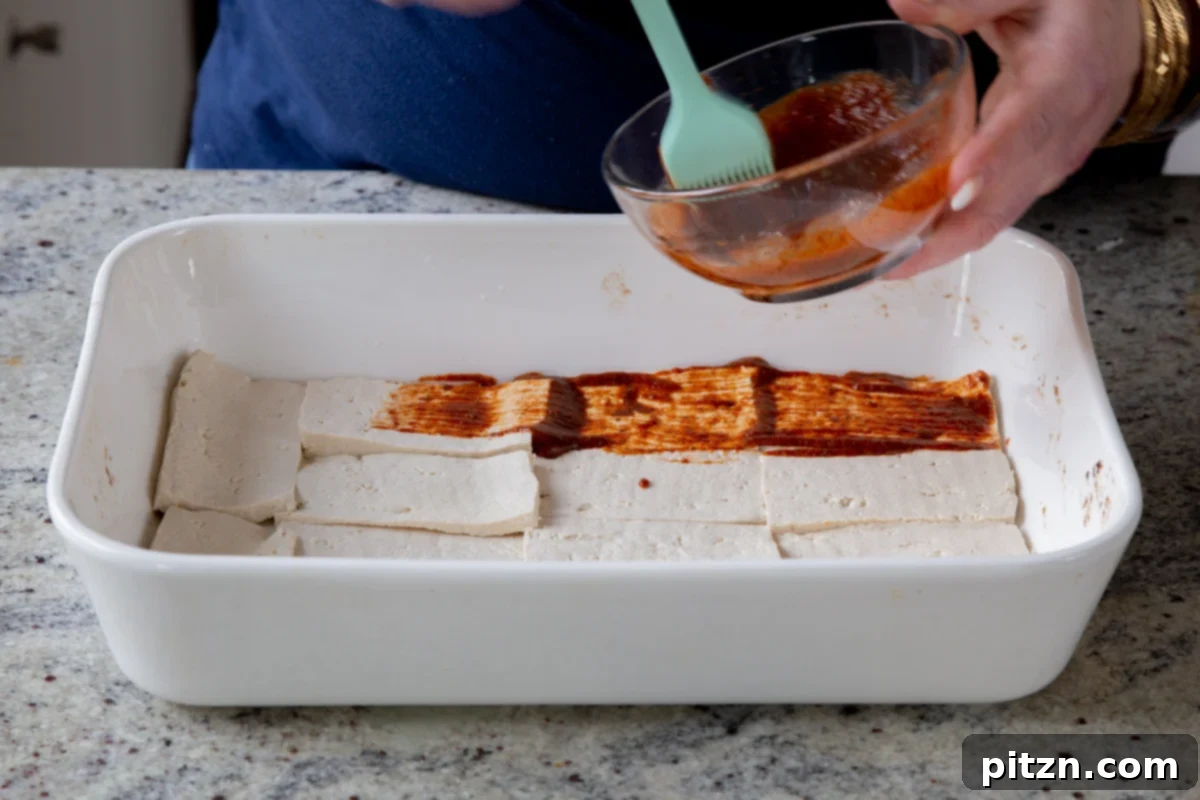 Plant-Based Deli Club 10 A brush expertly applying the rich marinade onto thin tofu deli slices arranged in a baking pan before baking.