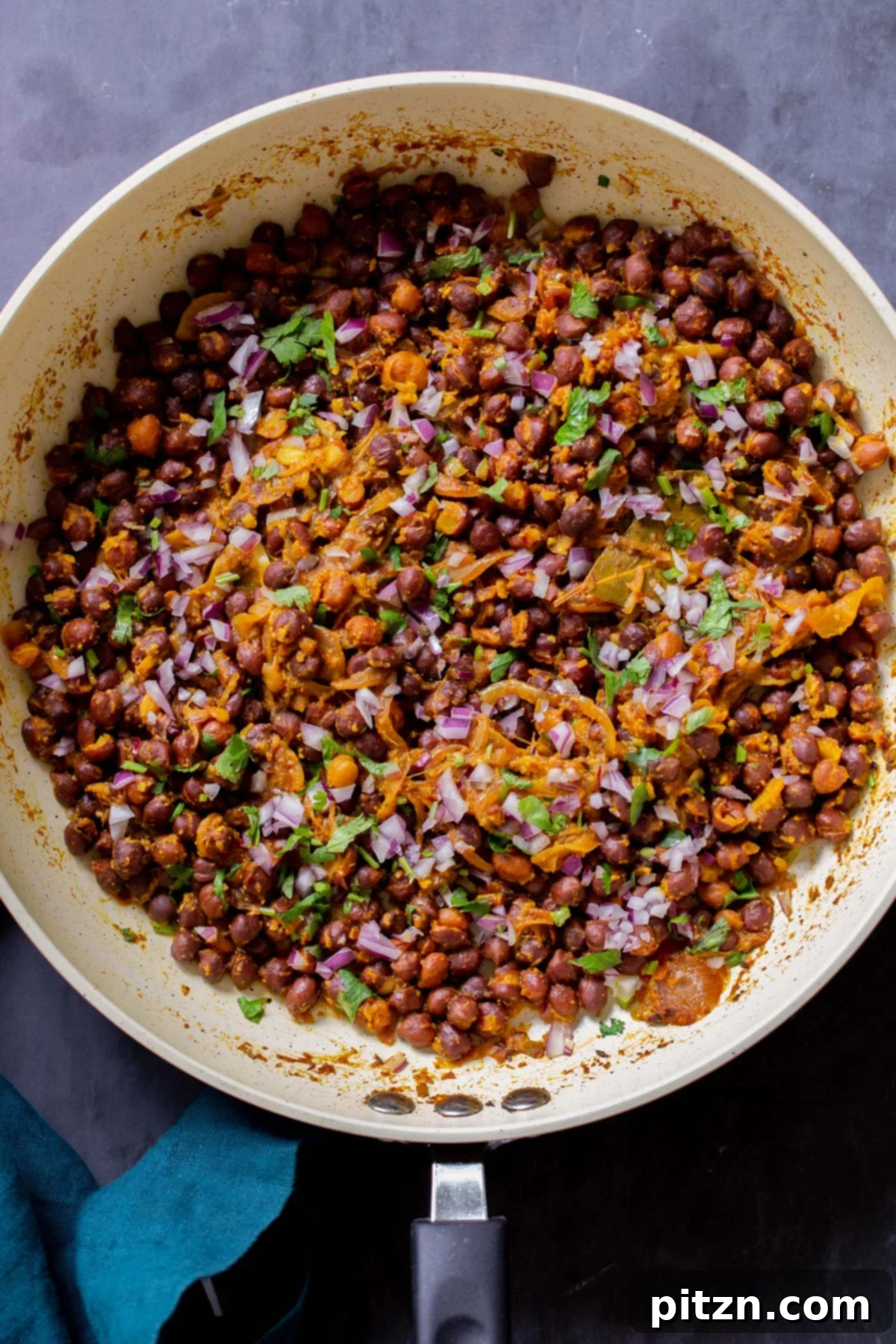 Flavorful Kala Chana Ghugni garnished with crunchy onions, fresh cilantro, and cumin, beautifully presented in a frying pan, ready to serve.