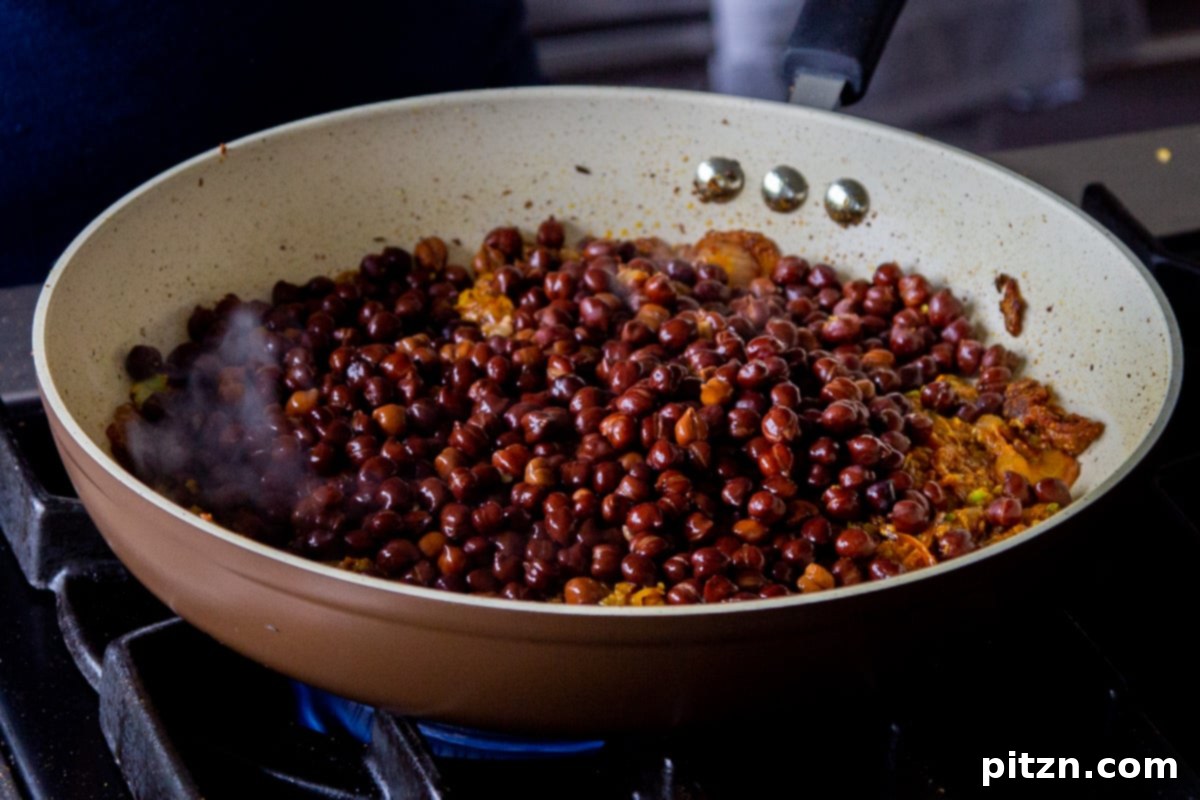 The step of adding the tender, cooked brown chickpeas to the vibrant, aromatic sauce simmering in the pan, ensuring all flavors combine perfectly.