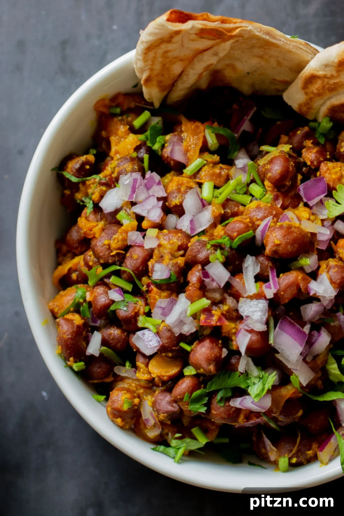 A close-up of a vibrant bowl of Kala Chana Ghugni, artfully garnished with a generous sprinkle of fresh cilantro, crunchy red onions, and aromatic toasted cumin.