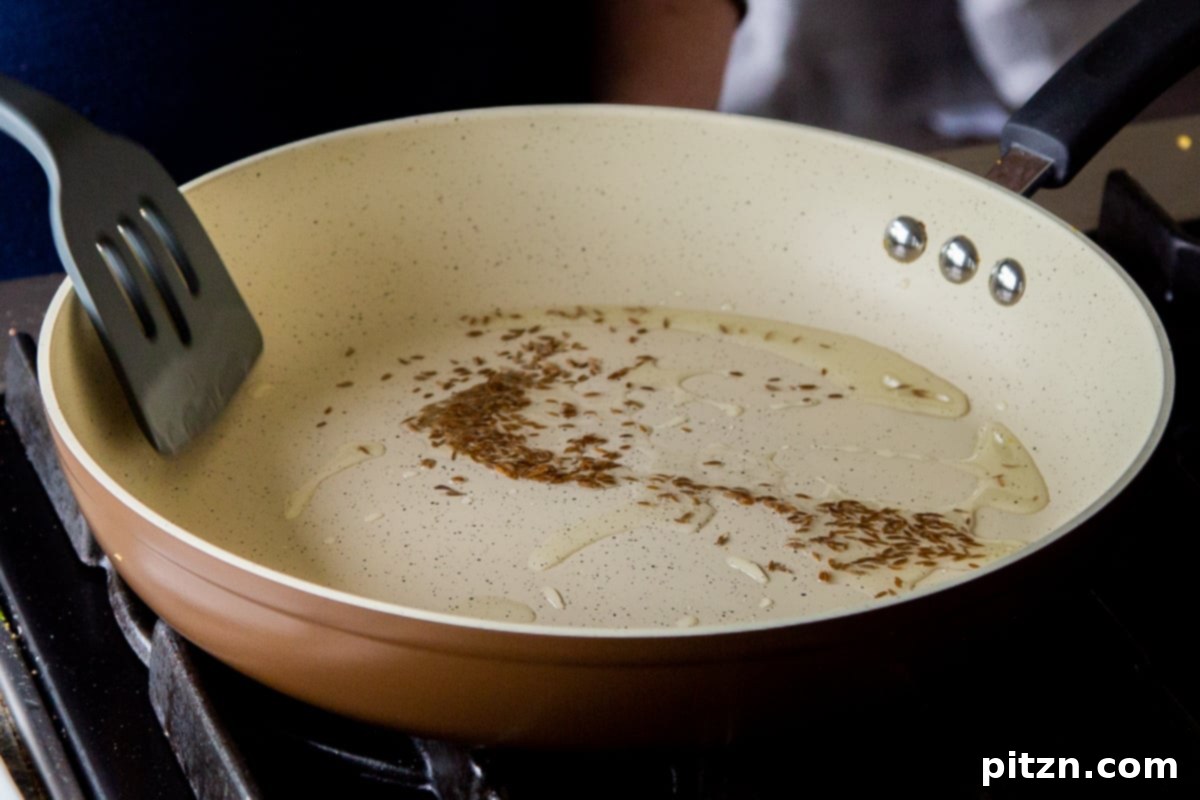 Whole cumin seeds sizzling and toasting in hot oil within a frying pan, indicating the start of the aromatic sauce preparation.