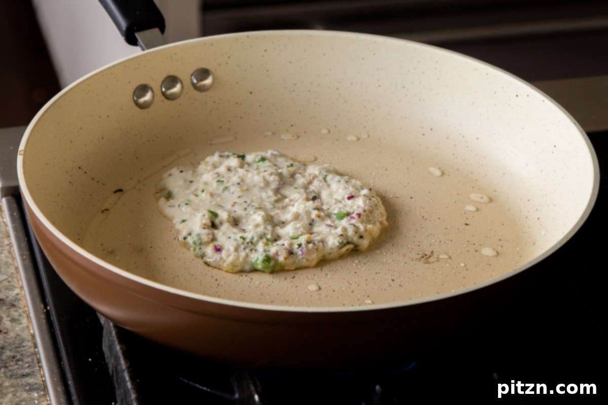 Raw Rava Idli pancake batter cooking in a hot skillet, forming a round, savory pancake before flipping.