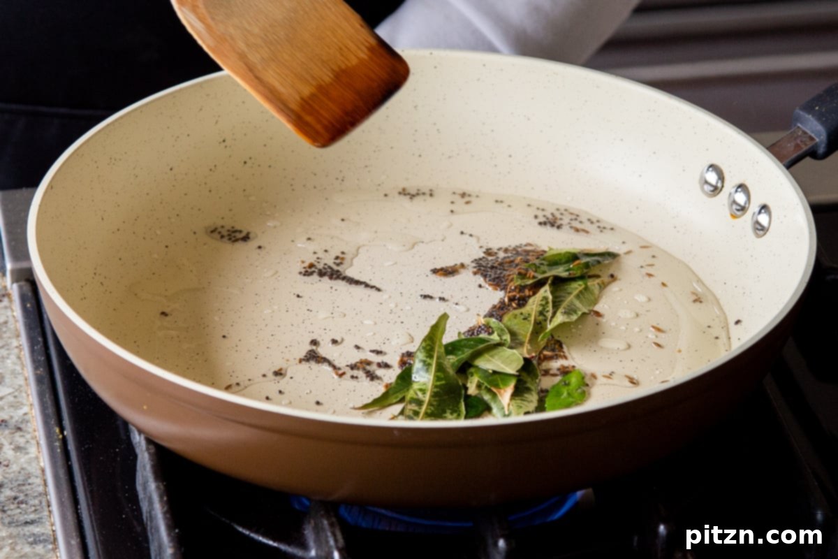 Adding fresh ginger, green chili, and curry leaves to the tempered whole spices in a hot pan for the Rava Idli Waffles batter.