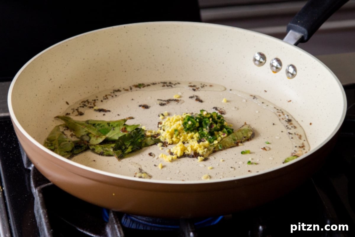 Chopped ginger and green chili being added to the pan with whole tempered spices, ready to infuse flavor into the Rava Idli Waffles batter.