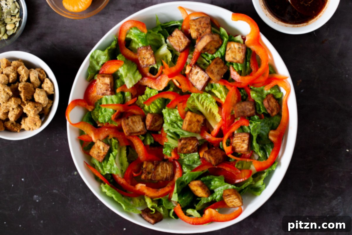 Smoky baked tofu pieces being added to the salad bowl with greens and bell pepper, providing a hearty protein element.
