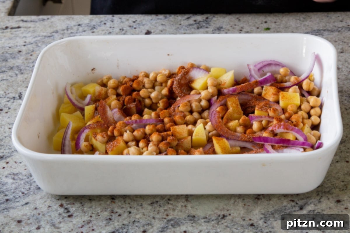 Smoky Chickpea Potato Salad 8 A hand sprinkling BBQ seasoning over chickpeas, onions, and potatoes in a baking dish, preparing them for roasting.