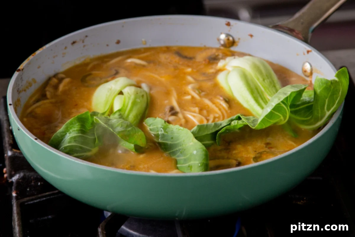 Adding fresh baby bok choy halves to the simmering Tonkotsu ramen, submerging them gently in the rich broth to cook until tender-crisp.