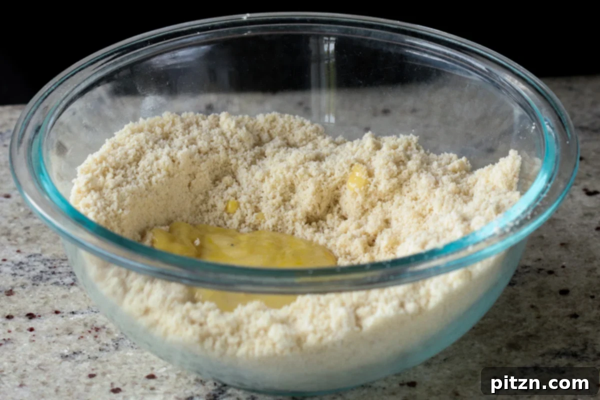 Adding a spoonful of saffron custard to the almond flour mixture in a bowl, beginning the process of forming the peda dough.