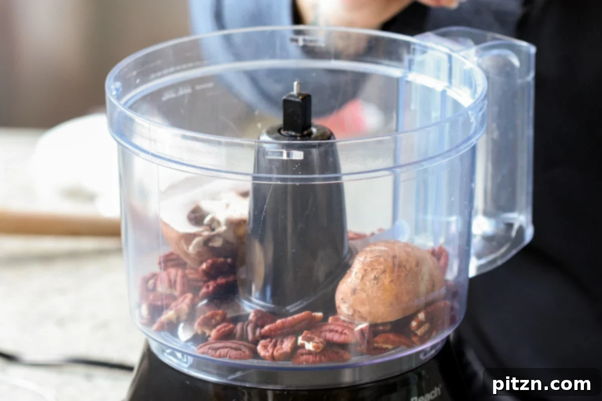 Pecans, chickpeas, and sliced mushrooms are clearly visible in the bowl of a food processor, positioned before the blending process begins.