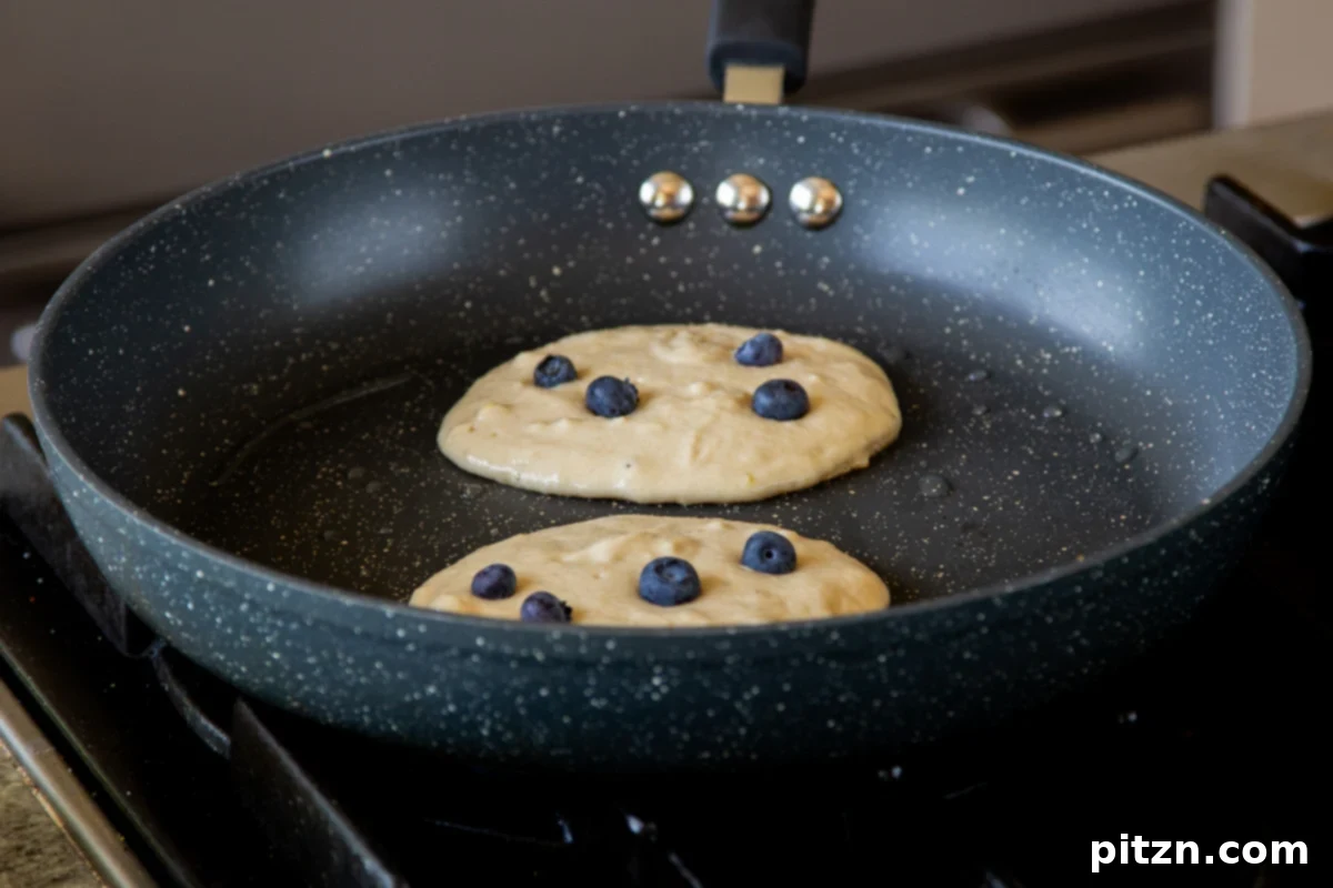 Gluten-free lemon blueberry pancakes cooking on a hot skillet, with visible blueberries embedded, before being flipped, showing their delicate rise