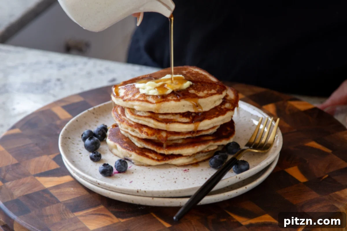A cascade of rich maple syrup being poured over a tall stack of golden gluten-free lemon blueberry pancakes, with fresh blueberries scattered around the plate, creating an enticing breakfast scene