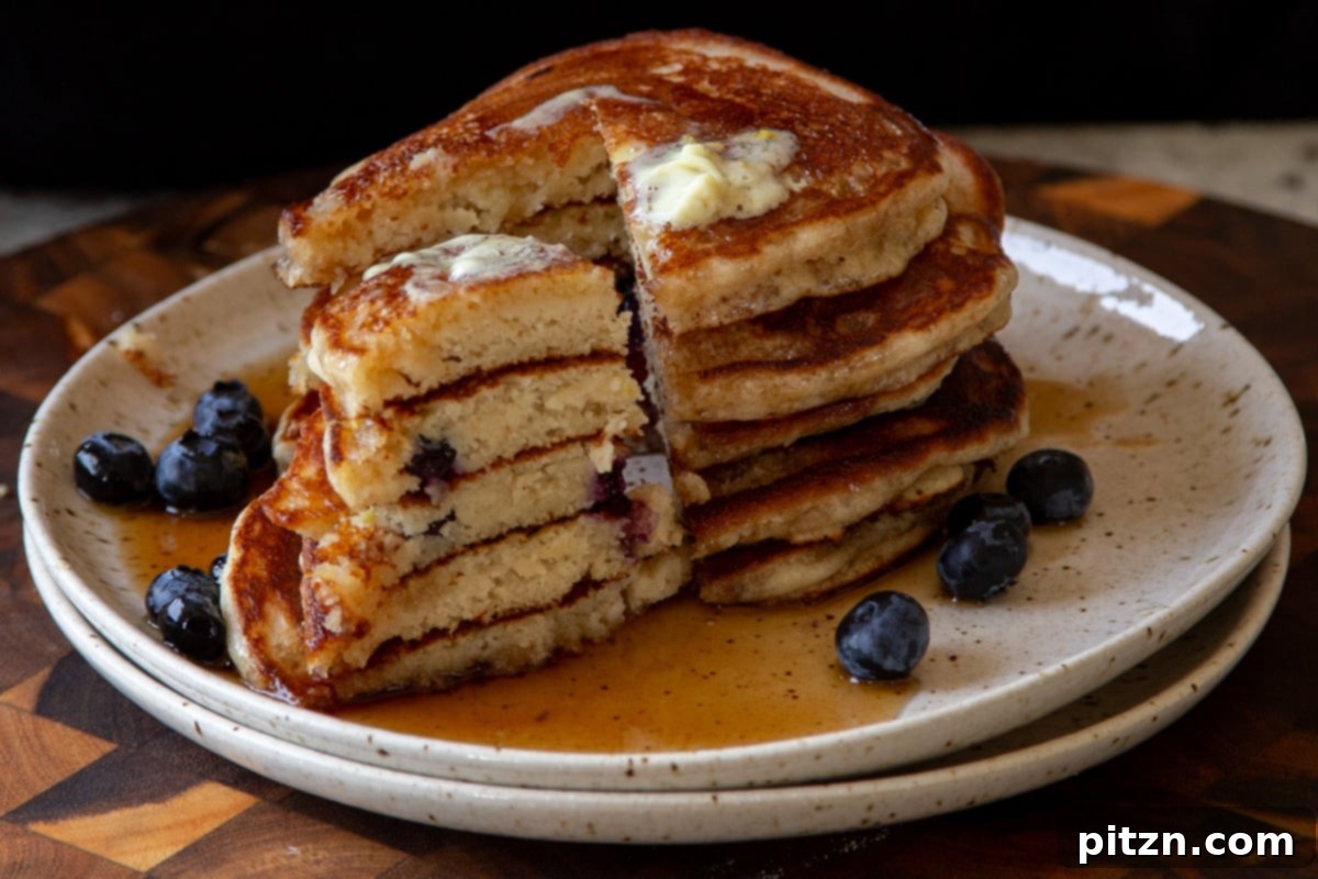 A stack of golden gluten-free lemon blueberry pancakes on a white plate, with a segment cut out to reveal the moist interior and bright blueberries, perfectly ready for a bite