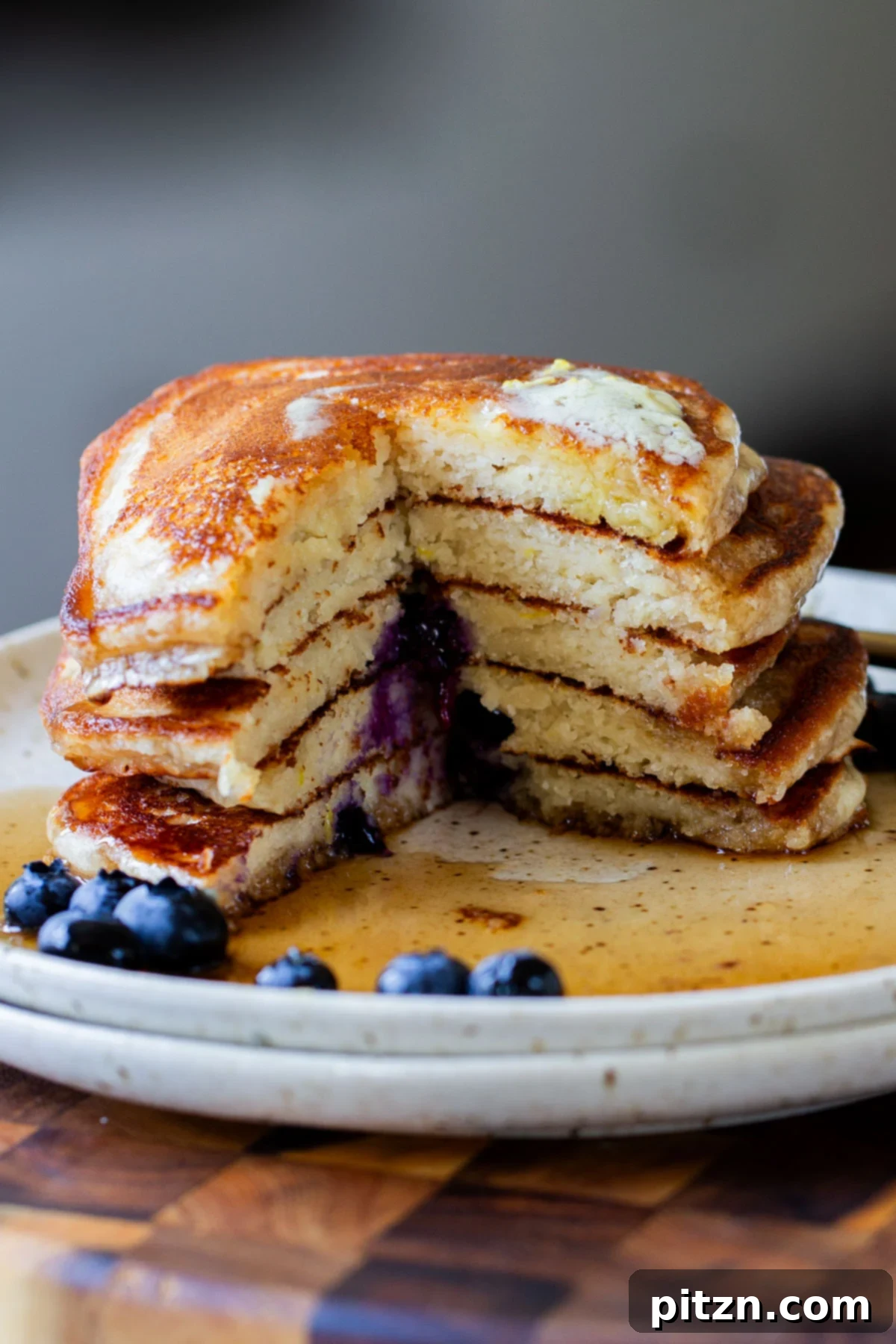 A close-up of a stacked gluten-free lemon blueberry pancake with a piece cut out, revealing its moist, tender interior and embedded blueberries, drizzled with maple syrup