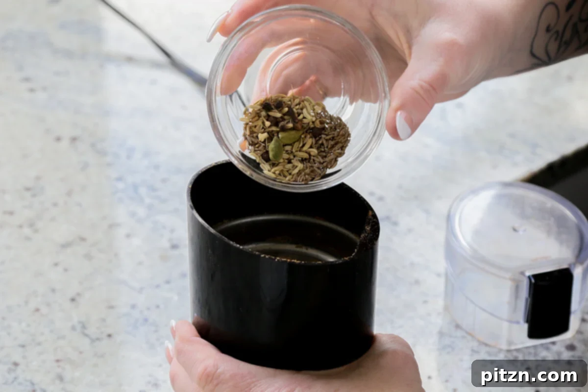 Fennel seeds, cumin seeds, cardamom pods, cloves, and black peppercorns are poured into a spice grinder to prepare the biryani's aromatic spice powder.