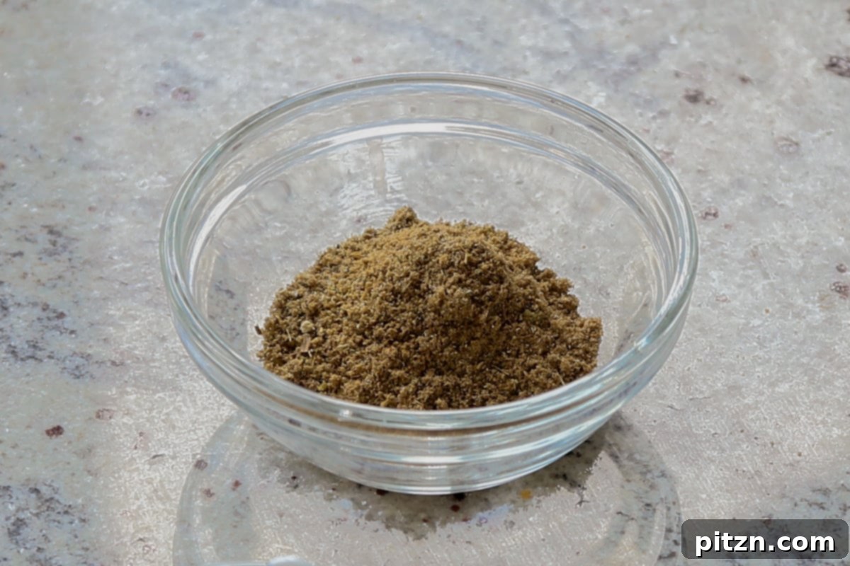 The freshly ground spice mix, a coarse powder of various whole and ground spices, rests in a small white bowl, radiating fragrance.