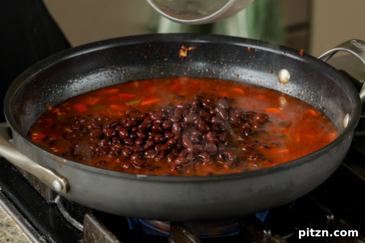 Adding black beans from a can to the pan with lentils and vegetables, completing the base for the burrito bowl.