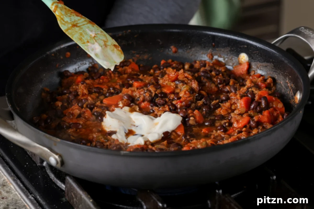 Stirring vegan sour cream into the fully cooked black bean and lentil mixture in the pan, adding creaminess and flavor.